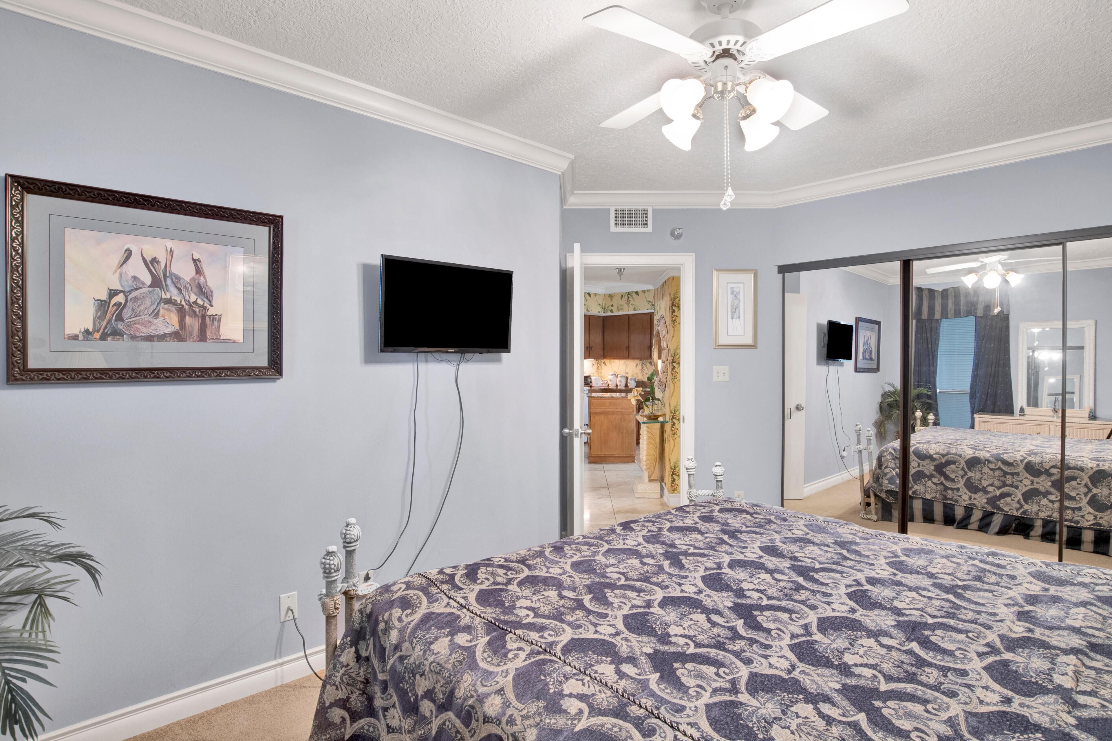 A bedroom with a bed, a television, a ceiling fan, a framed picture, and a door leading to a bathroom.