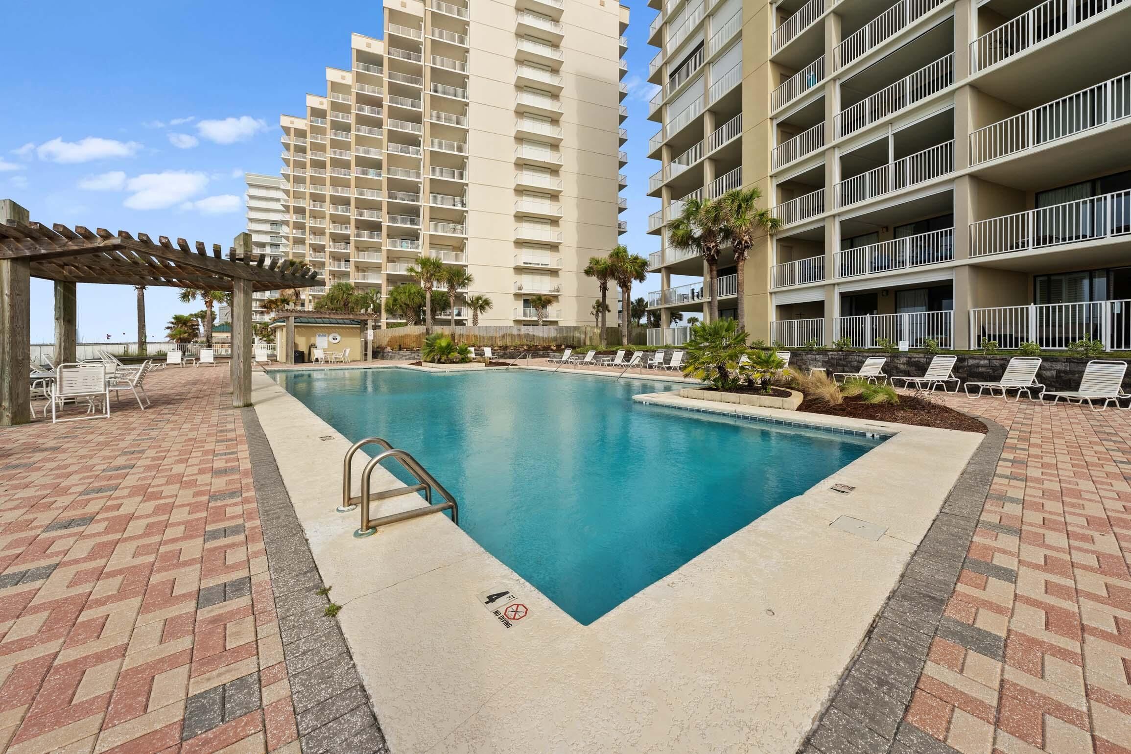 A swimming pool with a wooden deck and lounge chairs, surrounded by a high-rise building with balconies.