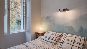 A bedroom with a bed, a nightstand with a lamp, a window with a view of trees, and a wall with a painted landscape. - La Marinière - Wifi - Bord de rivière (Laval)