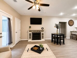 Una sala de estar con chimenea, ventilador de techo, comedor y cocina visible a través de un ventanal.