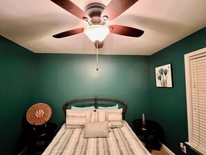 Een slaapkamer met een bed, een plafondventilator, een nachtkastje en een aan de muur bevestigd schilderij.