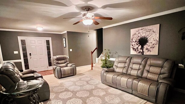 A living room with a gray sofa, a recliner, a glass coffee table, a patterned rug, a ceiling fan, and a staircase.