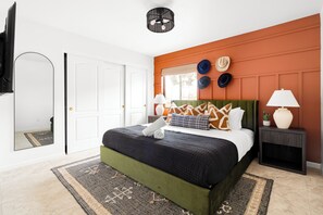 Ein Schlafzimmer mit einem grünen, gepolsterten Bett, einer weißen Tür, einem Spiegel und einem an der Wand montierten Fernseher.