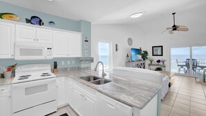 A modern kitchen with white cabinets, a built-in stove, and a large island with a sink. There is a view of the ocean through a large window, and a ceiling fan is visible. - Tropic Isles 801 (Gulf Shores)