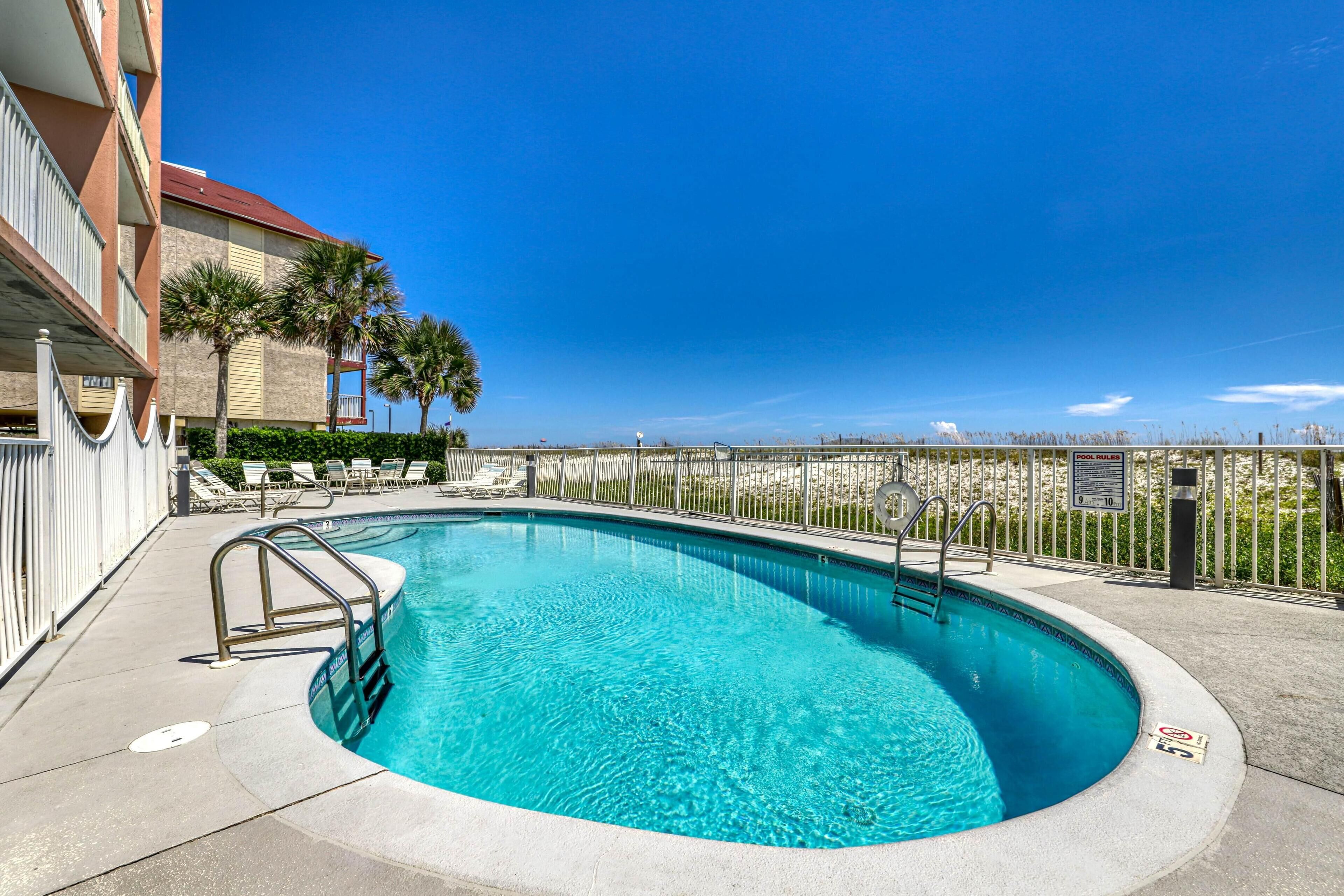 A swimming pool with a metal ladder, surrounded by a fence, and a view of a building with balconies and palm trees.