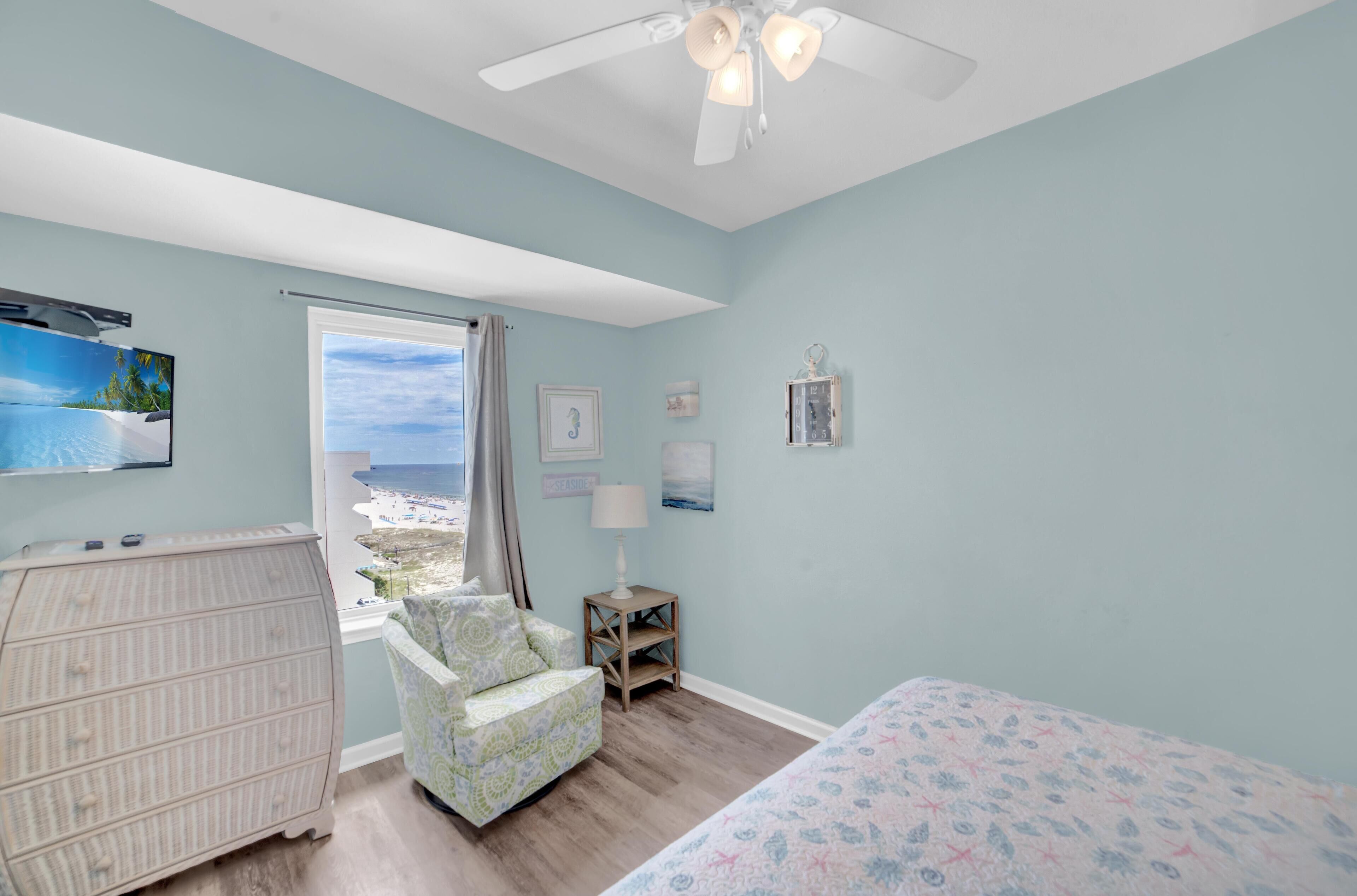 A bedroom with a bed, a chair, a dresser, a window with a view of the beach, and a ceiling fan.
