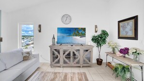 A living room with a large TV, a clock, a window with a view, a sofa, and a cabinet with decorative items. - Tropic Isles 801 (Gulf Shores)