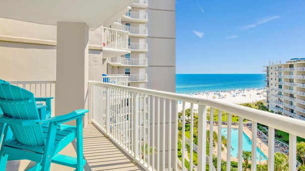 Een balkon met turquoise stoelen, een witte balustrade en uitzicht op een strand en zwembad.