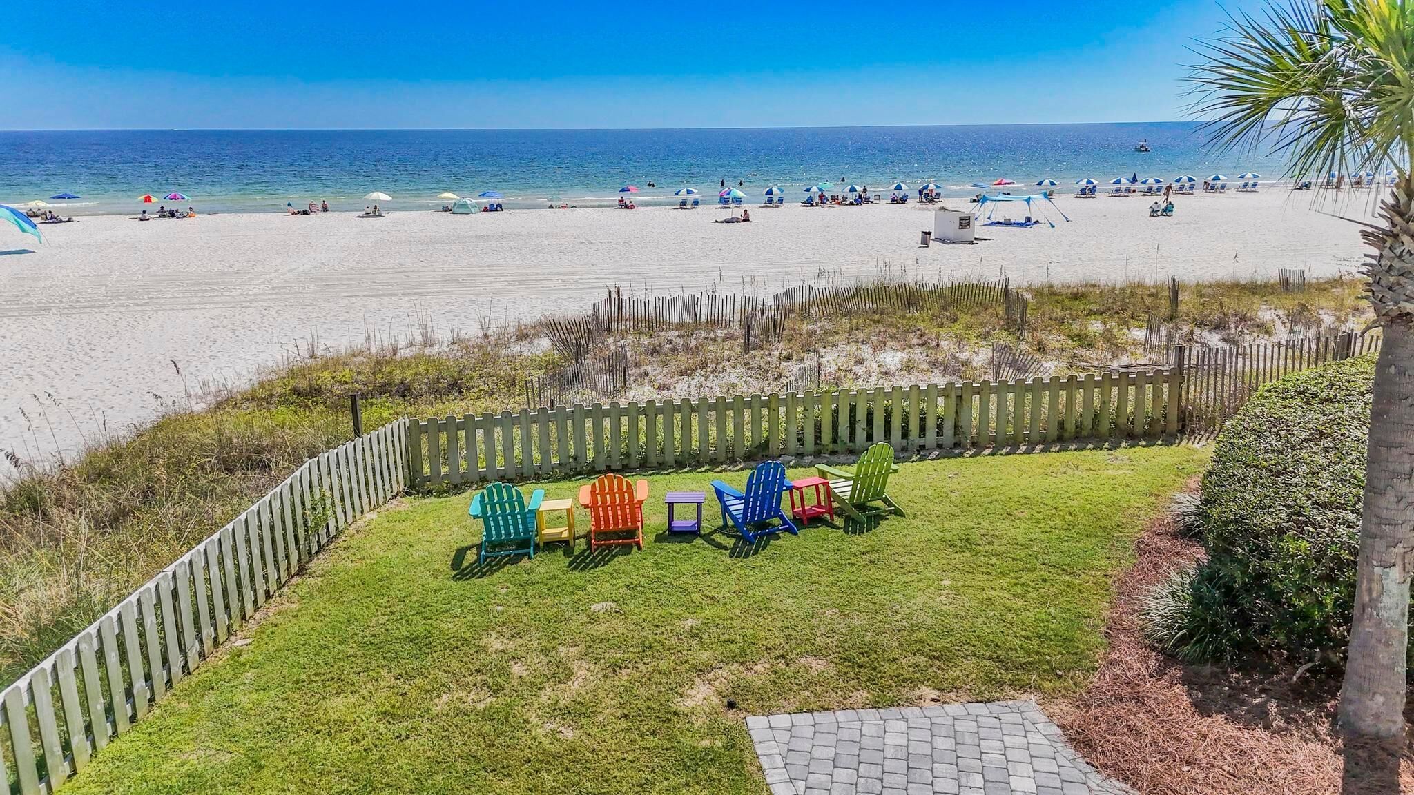 A beach view with a grassy area and colorful Adirondack chairs.