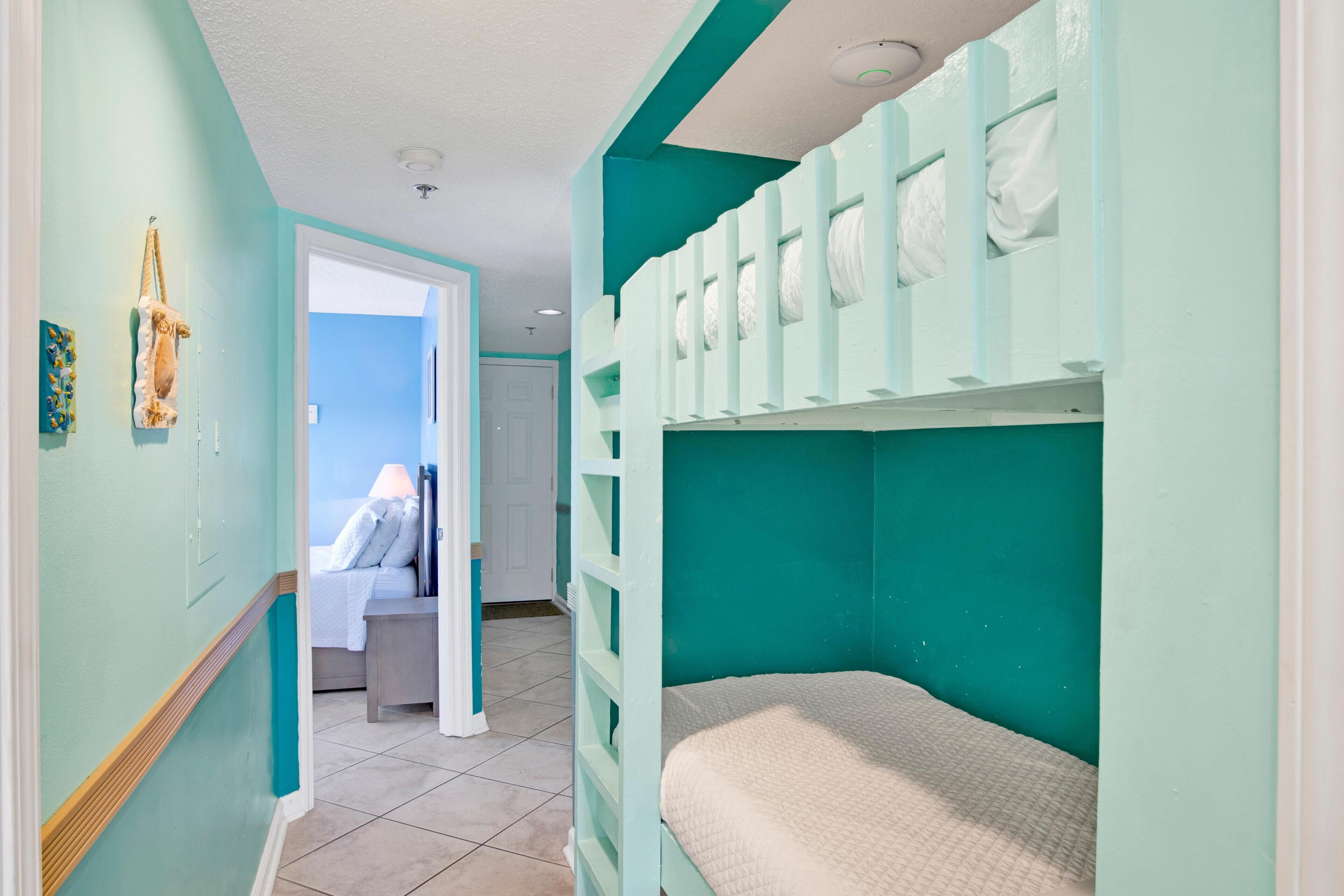 A room with teal walls, a bunk bed, a bedside table, and a door leading to another room.