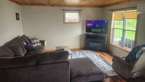Uma sala de estar com um sofá marrom escuro, uma poltrona pequena, uma TV de tela plana em um suporte e uma janela com persianas.
