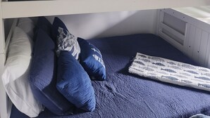 Quarto com beliche de dois andares, cada um com colchão e almofadas. Há uma janela com cortinas e um ventilador de teto à vista.