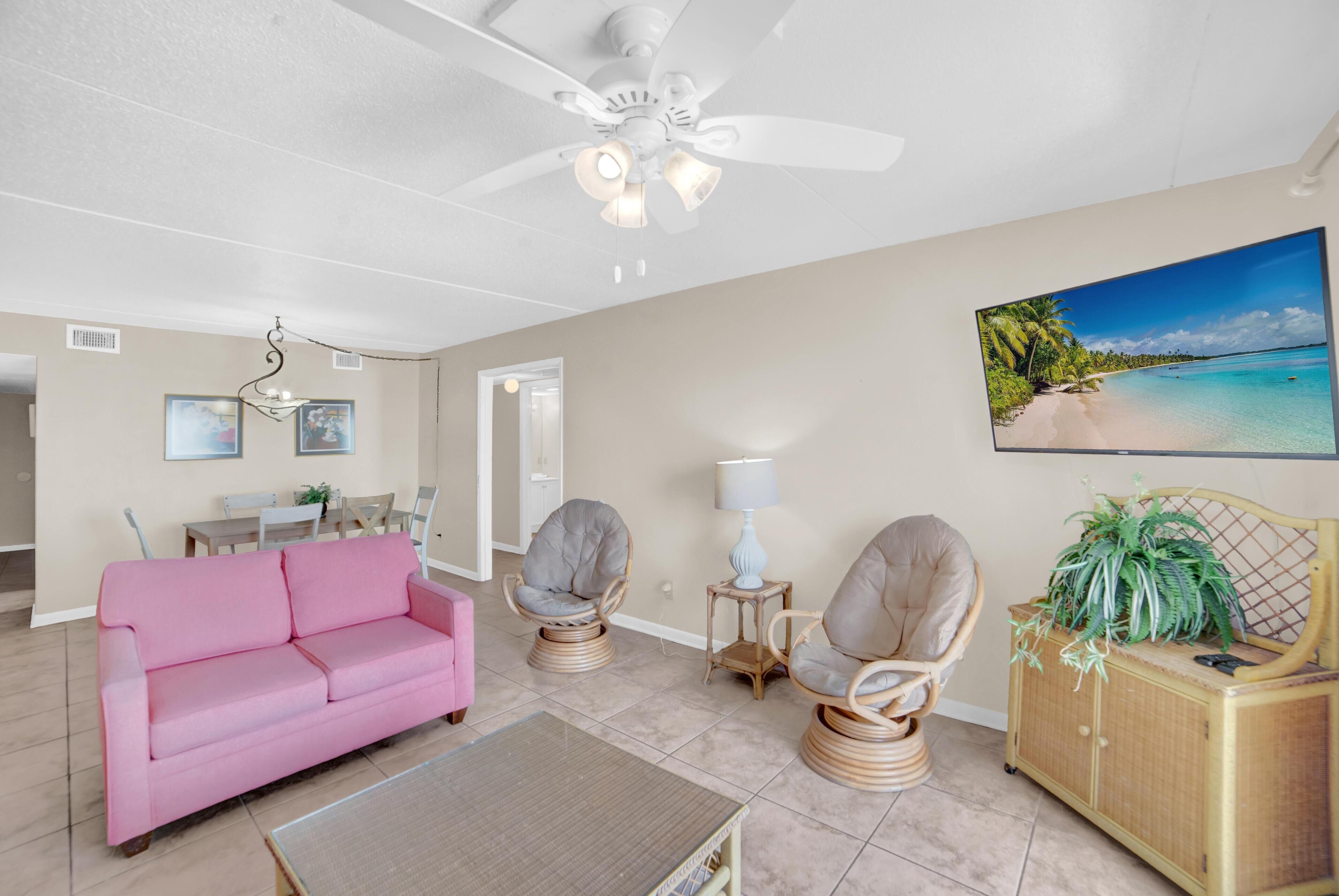 A living room with a pink sofa, wicker chairs, a coffee table, a ceiling fan, and a beach-themed painting.