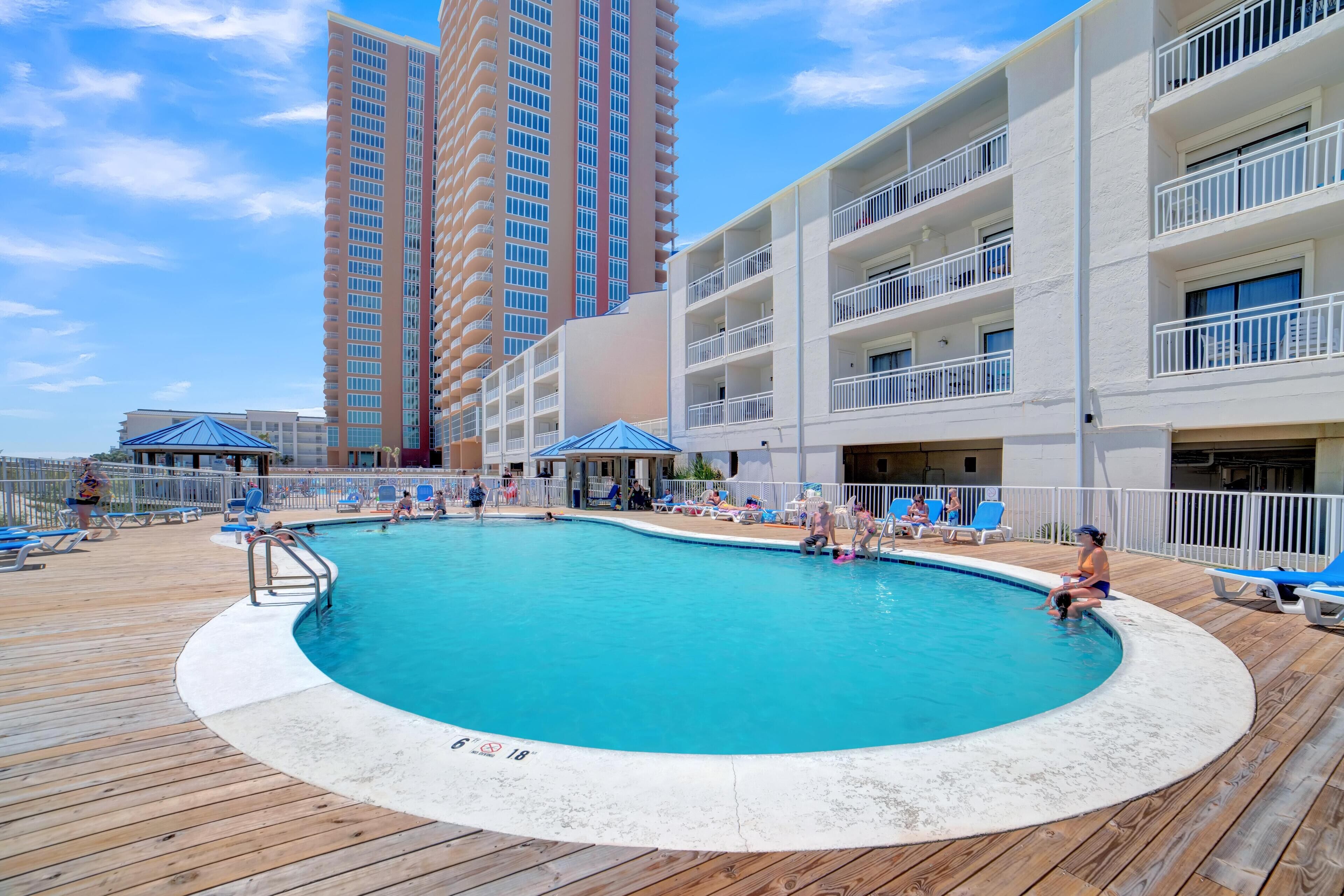A pool area with a wooden deck, lounge chairs, and a view of tall buildings in the background.