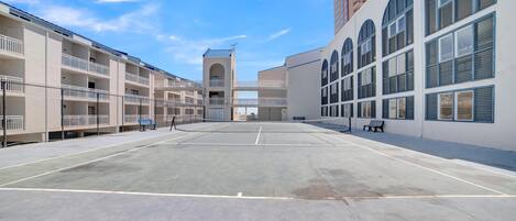 A tennis court with a net in the center, surrounded by buildings with balconies and large windows.