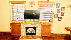 Ein Wohnzimmer mit Kamin aus Stein, einem flachbildfernseher darüber, Holzjalousien an den Fenstern und mehreren gerahmten Bildern an der Wand.