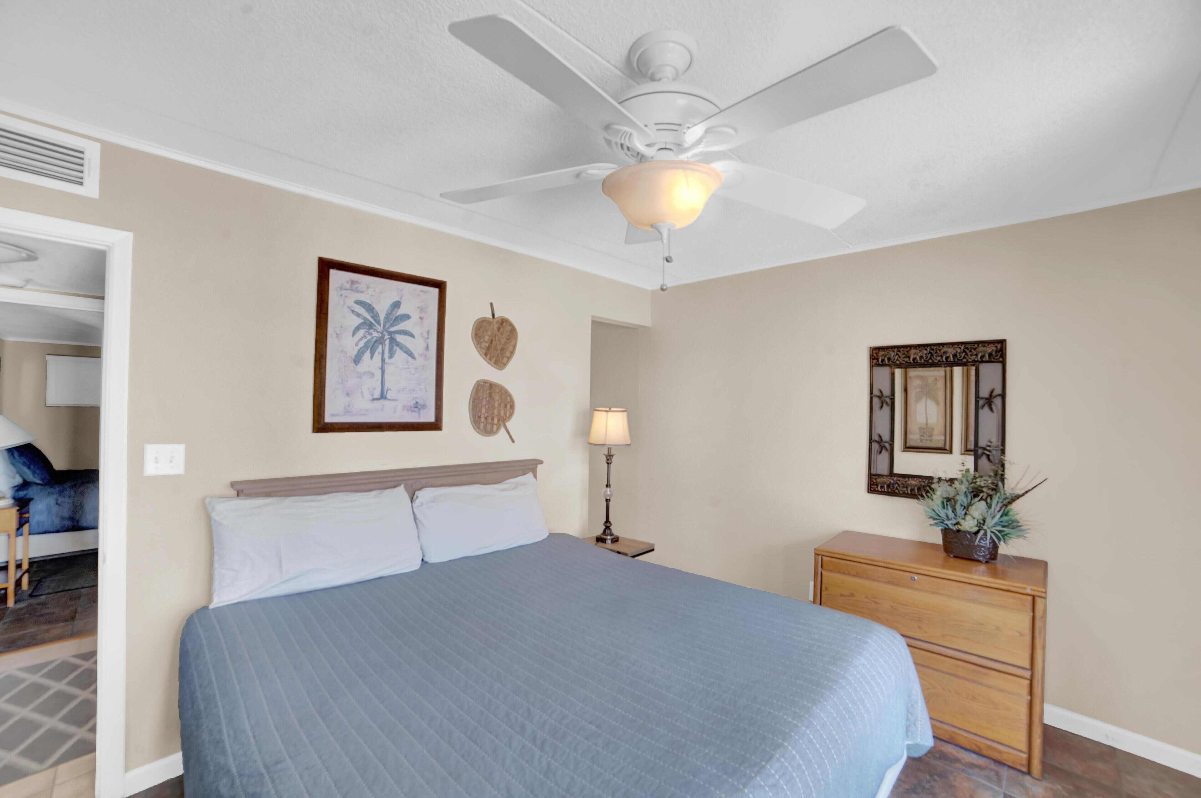 A bedroom with a bed, a ceiling fan, a nightstand with a lamp, a dresser, and wall decorations including framed pictures and a mirror.