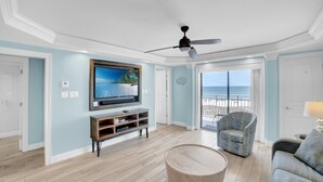 A spacious living room with a large flat-screen TV, a ceiling fan, a wooden coffee table, a patterned armchair, and a view of the ocean through a sliding glass door. - Sugar Beach 241 (Orange Beach)