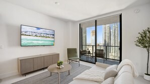 A modern living room with a large TV, a sofa, a chair, a small table, and a view of tall buildings through large windows. - 601 RESIDENCES SKY FLOOR 1BR UNIT #2611 (Miami)