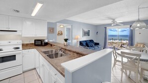 A modern kitchen with white cabinets, a granite countertop, and stainless steel appliances. There is a dining area with white chairs and a table, and a balcony with a view of the ocean. - Sugar Beach 245 (Orange Beach)