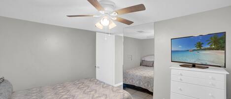 A bedroom with a ceiling fan, a bed with a patterned bedspread, a dresser with a television displaying a beach scene, and a door leading to another room.