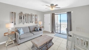 A living room with a grey sofa, a wooden coffee table, and a ceiling fan. There is a view of the ocean through a sliding glass door. - Sugar Beach 246 (Orange Beach)