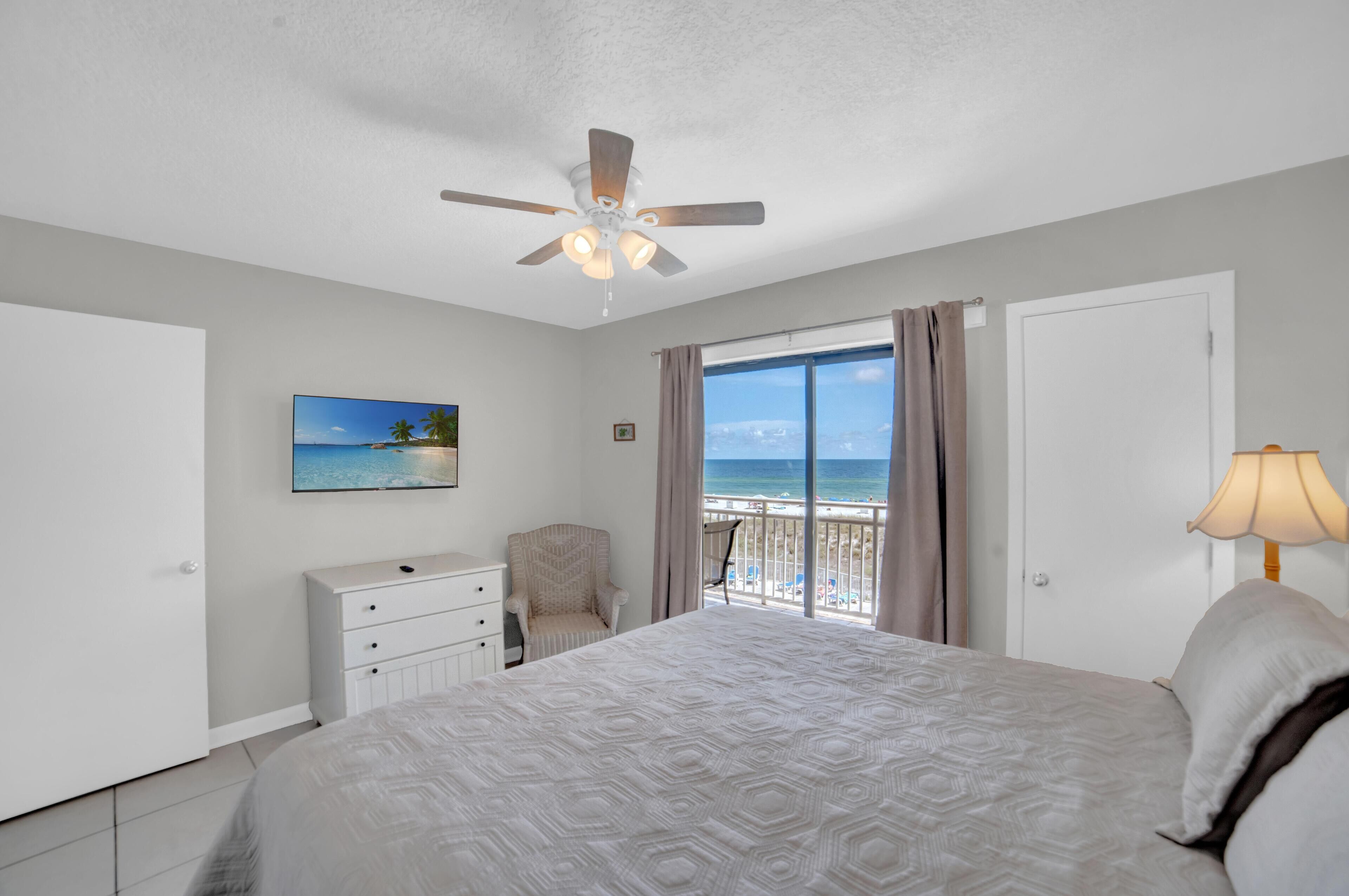 A bedroom with a large bed, a ceiling fan, a nightstand, a chair, a dresser, a window with a view of the beach, and a lamp.