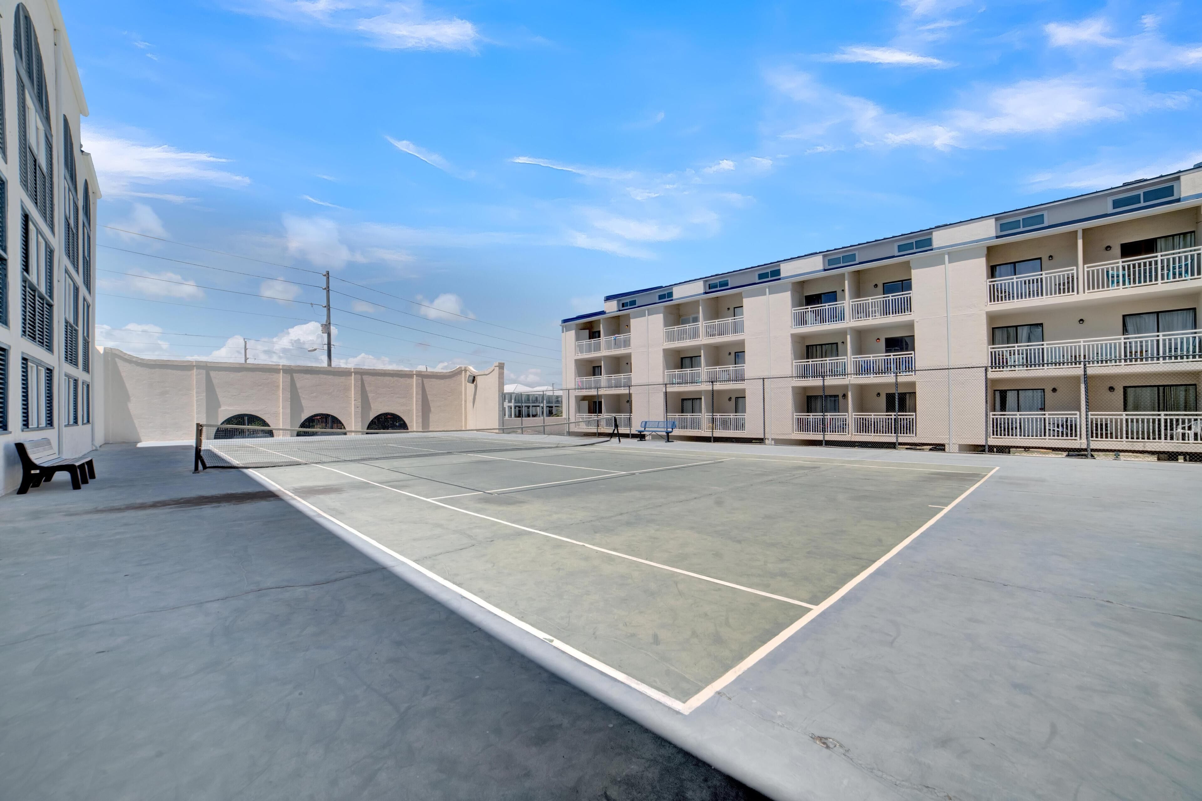 An outdoor tennis court with a net, surrounded by a building with balconies and a clear sky.
