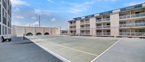 An outdoor tennis court with a net, surrounded by a building with balconies and a clear sky.