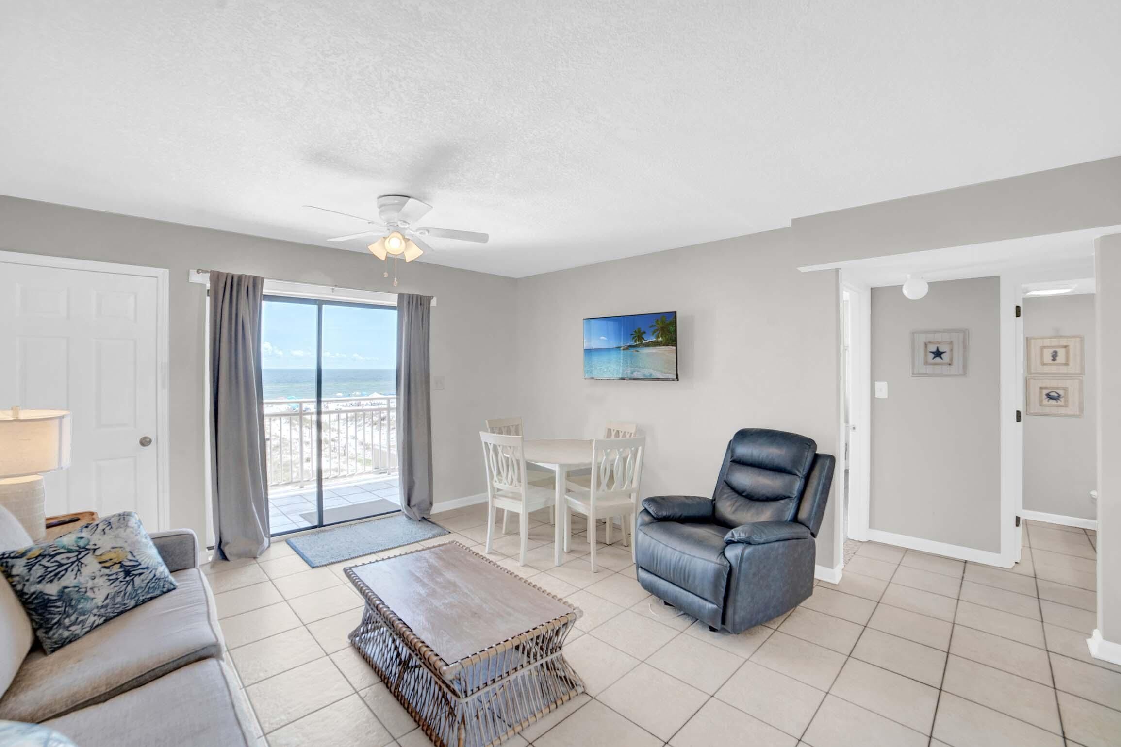 Un salon comprenant un canapé, une table basse, une table à manger avec des chaises, un ventilateur de plafond et un tableau représentant une scène de plage.