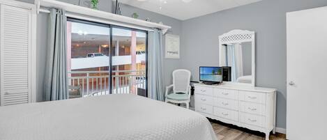 A bedroom with a large bed, a ceiling fan, a TV, a dresser, and a balcony with a view of the street.