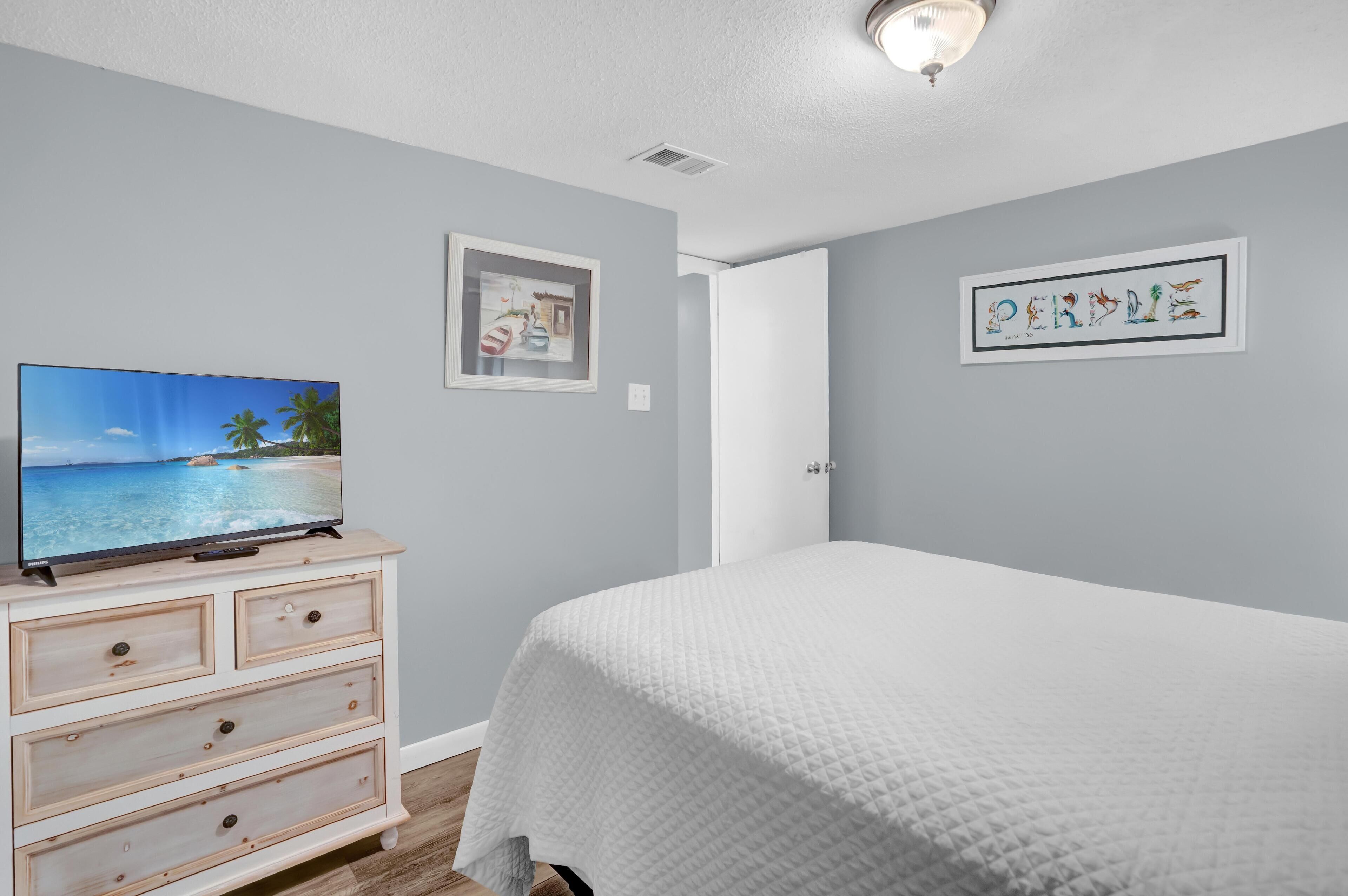 Un dormitorio con una cama, un aparador, un televisor mostrando una escena de playa y dos cuadros en la pared.