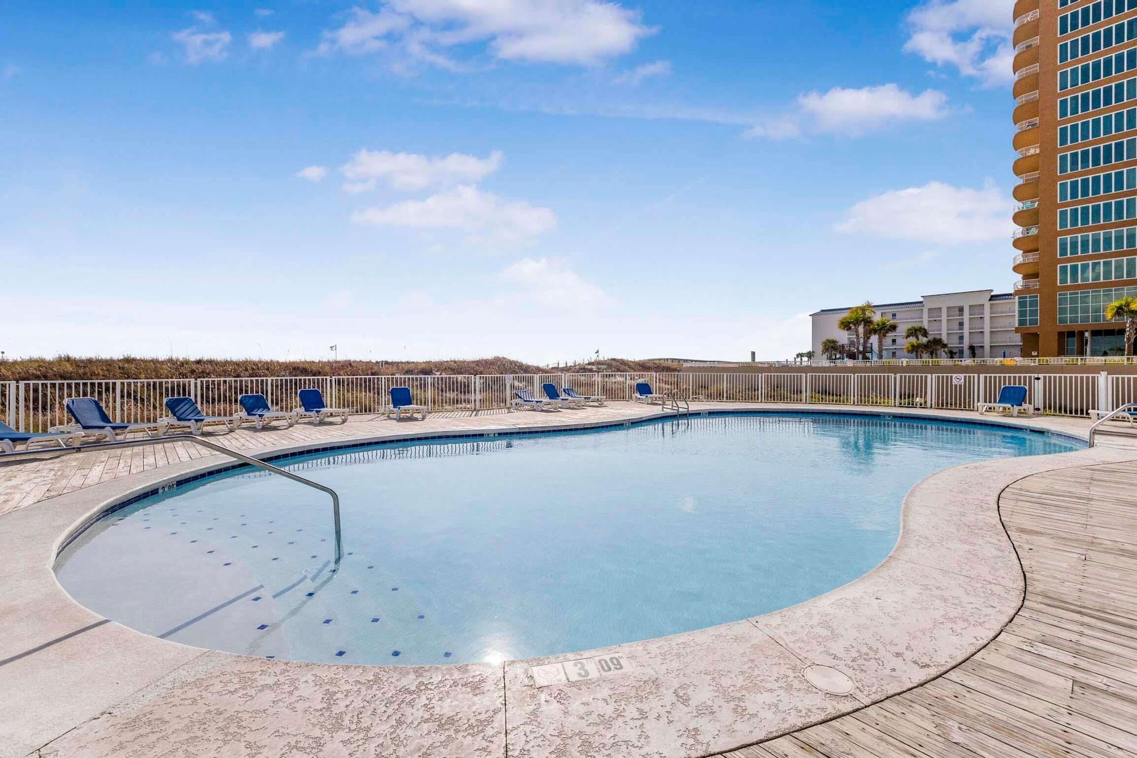 A swimming pool with a wooden deck and lounge chairs, surrounded by a fence and a building with balconies.