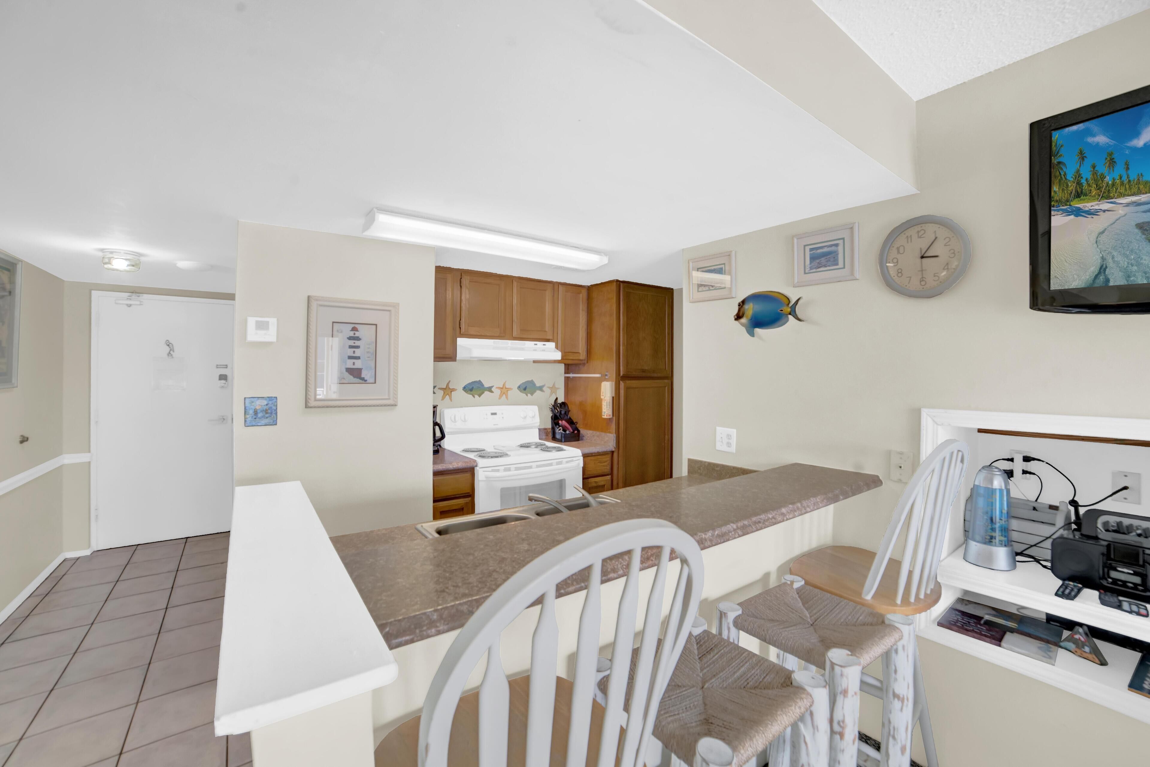 A compact kitchen with a white stove, wooden cabinets, and a white countertop. There are two white chairs and a small table.