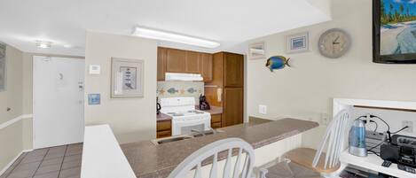 A compact kitchen with a white stove, wooden cabinets, and a white countertop. There are two white chairs and a small table.