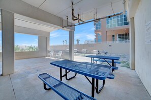 An outdoor patio area with blue picnic tables and chairs, concrete pillars, and a view of a building with a sign that reads "Manshield." - Sugar Beach 323 (Orange Beach)