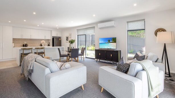 Una sala moderna con un sofá gris, un comedor con mesa y sillas, una cocina con armarios blancos y un ventanal con vistas al exterior.