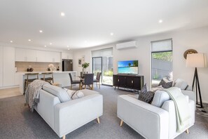 Ein modernes Wohnzimmer mit einem grauen Sofa, einem Essbereich mit Tisch und Stühlen, einer Küche mit weißen Schränken und einem großen Fenster mit Blick ins Freie.