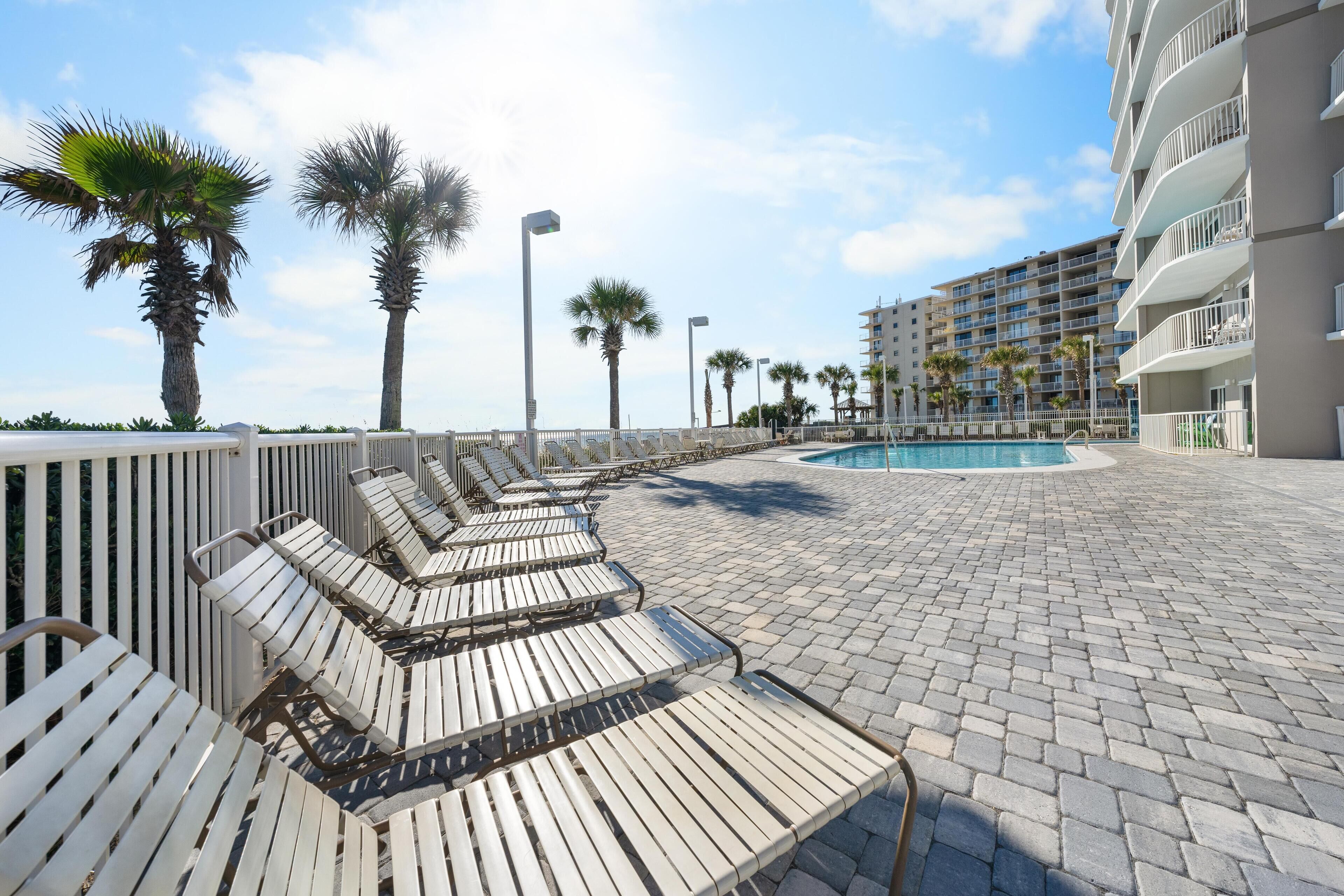 A poolside area with lounge chairs, palm trees, and a building in the background.