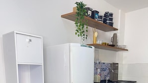 Cocina compacta con encimera blanca, dos taburetes negros, un refrigerador blanco, una estufa y estantes con utensilios de cocina y una planta en maceta.