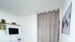 Una sala de estar con un sofá gris, un televisor sobre un mueble blanco, un armario blanco y una cortina.