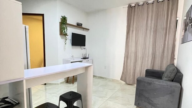 Un espacio habitable compacto con kitchenette, una zona de comedor con dos taburetes, un sofá gris y un televisor montado en la pared.