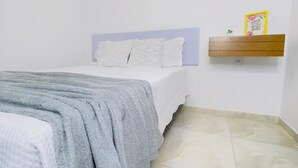Un dormitorio con una cama, una manta gris, un edredón blanco, un estante de madera con una planta pequeña y un cuadro colgado en la pared.