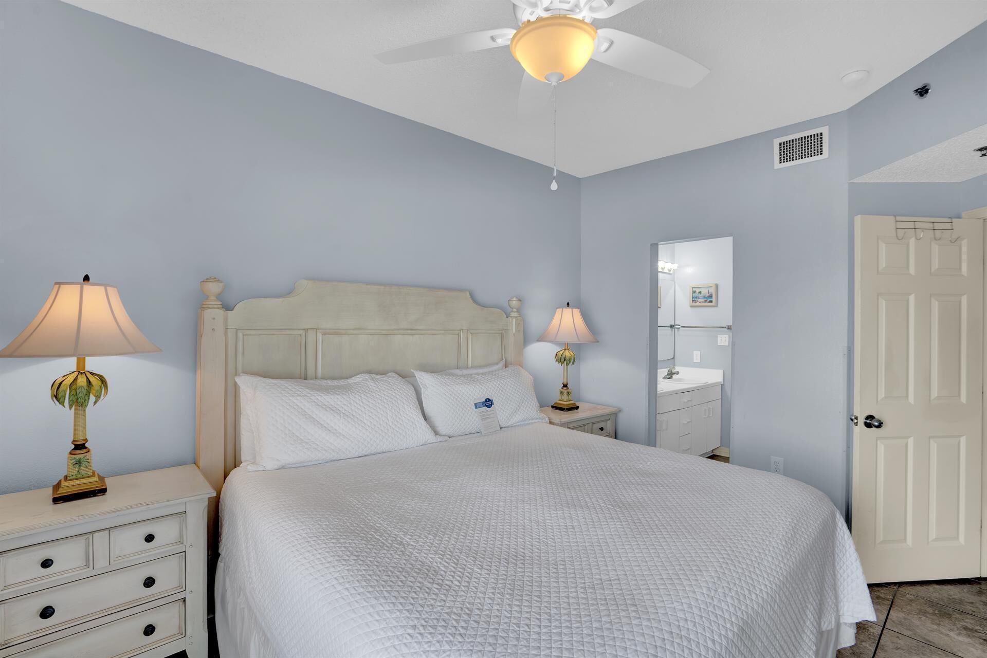 A bedroom with a bed, two bedside tables with lamps, a ceiling fan, a bathroom with a sink and mirror, and a door.