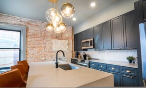 A modern kitchen with a brick wall, dark cabinets, a large island with a sink, and pendant lighting. - The Buckeye Retreat Mansion (Columbus)