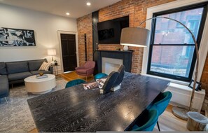 Un salon moderne avec un mur en briques, une cheminée, une table à manger avec des chaises, un canapé et un téléviseur fixé au mur.