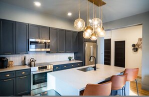 Une cuisine moderne avec des meubles de couleur sombre, des appareils en acier inoxydable, un grand îlot central et des luminaires suspendus.