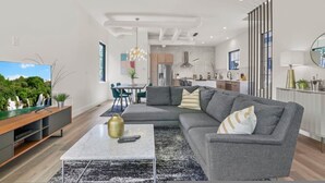 Un salon moderne avec un canapé d’angle, une table basse et un téléviseur. On y trouve également un coin repas avec une table et des chaises, et une cuisine à l’arrière-plan.