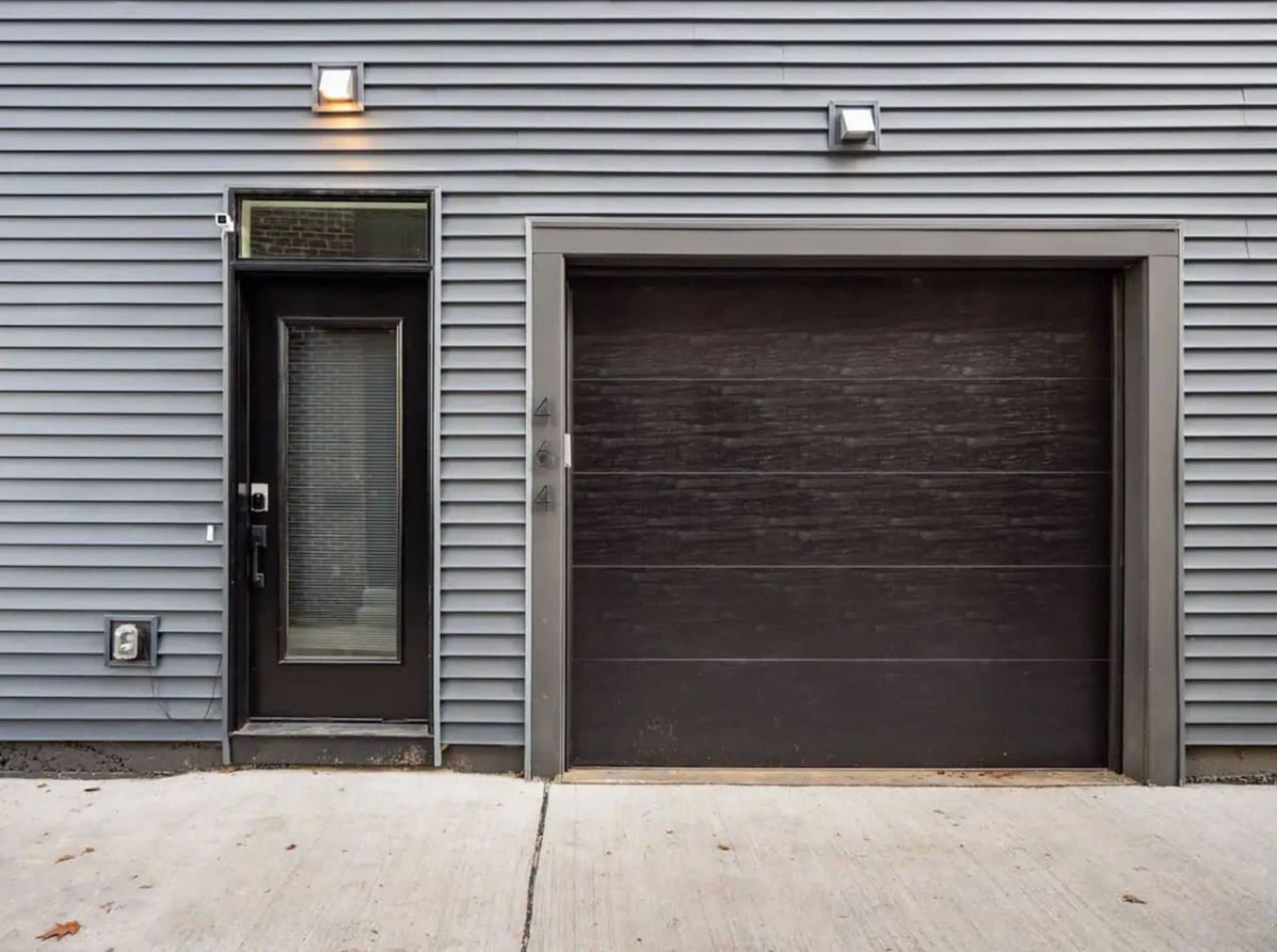 Une porte de garage à la finition sombre, une porte avec un panneau en verre et un petit luminaire placé au-dessus de la porte.