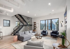 A modern living room with a grey sectional sofa, a coffee table, two armchairs, a staircase, and large windows providing a view of the outside. - Historic German Village Industrial Masterpiece (Columbus)